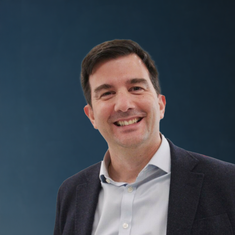A man with short dark hair, smiling, wearing a white shirt and dark blazer, standing against a dark blue background.