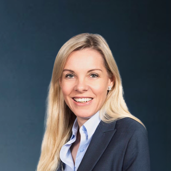 Portrait of a smiling woman with long blonde hair wearing a navy blazer and light blue collared shirt against a dark background.