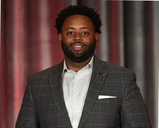 A man with black curly hair and a beard wearing a gray plaid blazer over a white shirt, standing in front of a red curtain background.