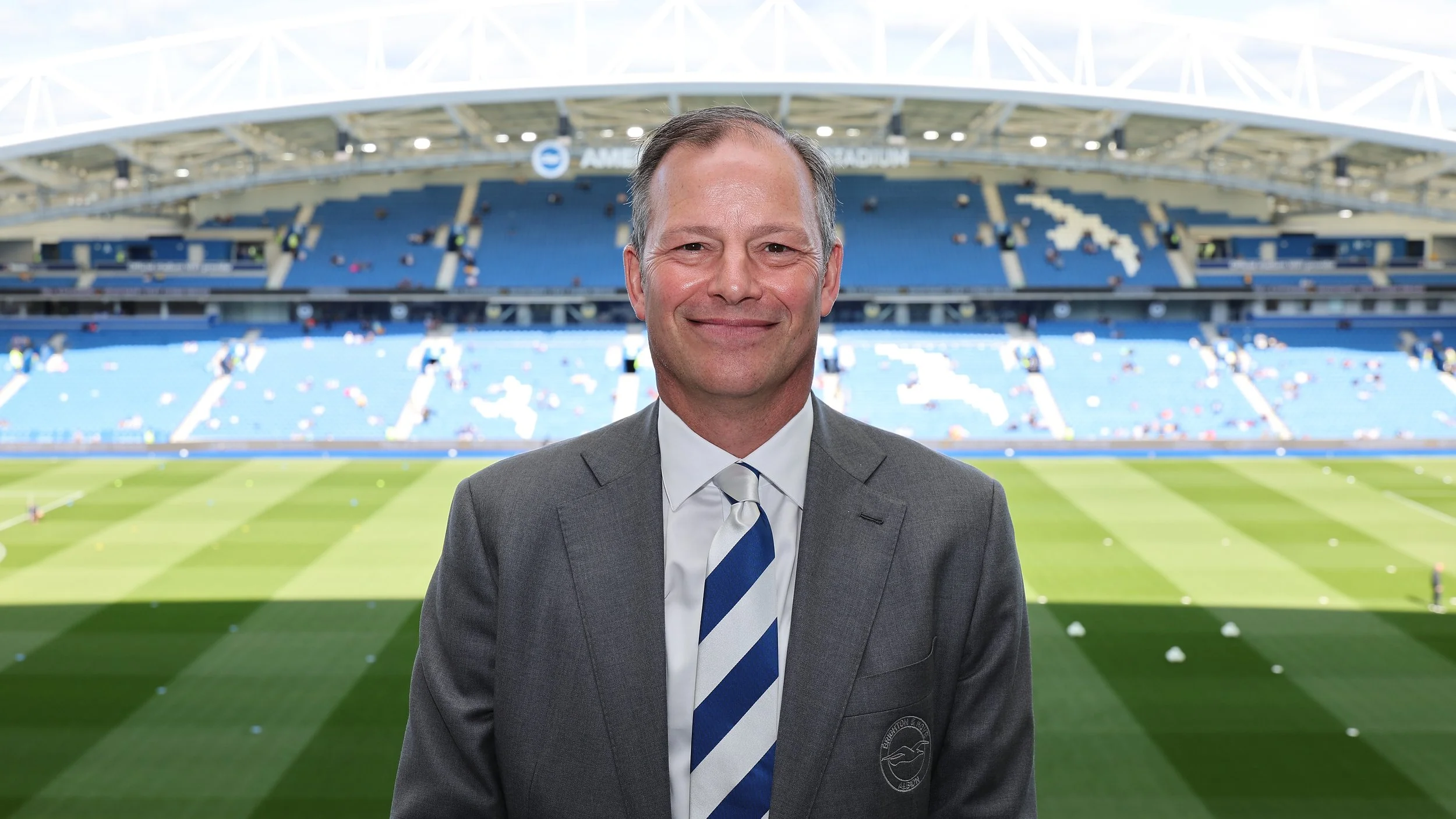 A smiling man in a suit standing on a football stadium field, with empty blue seats and a partial view of the stadium's roof in the background.