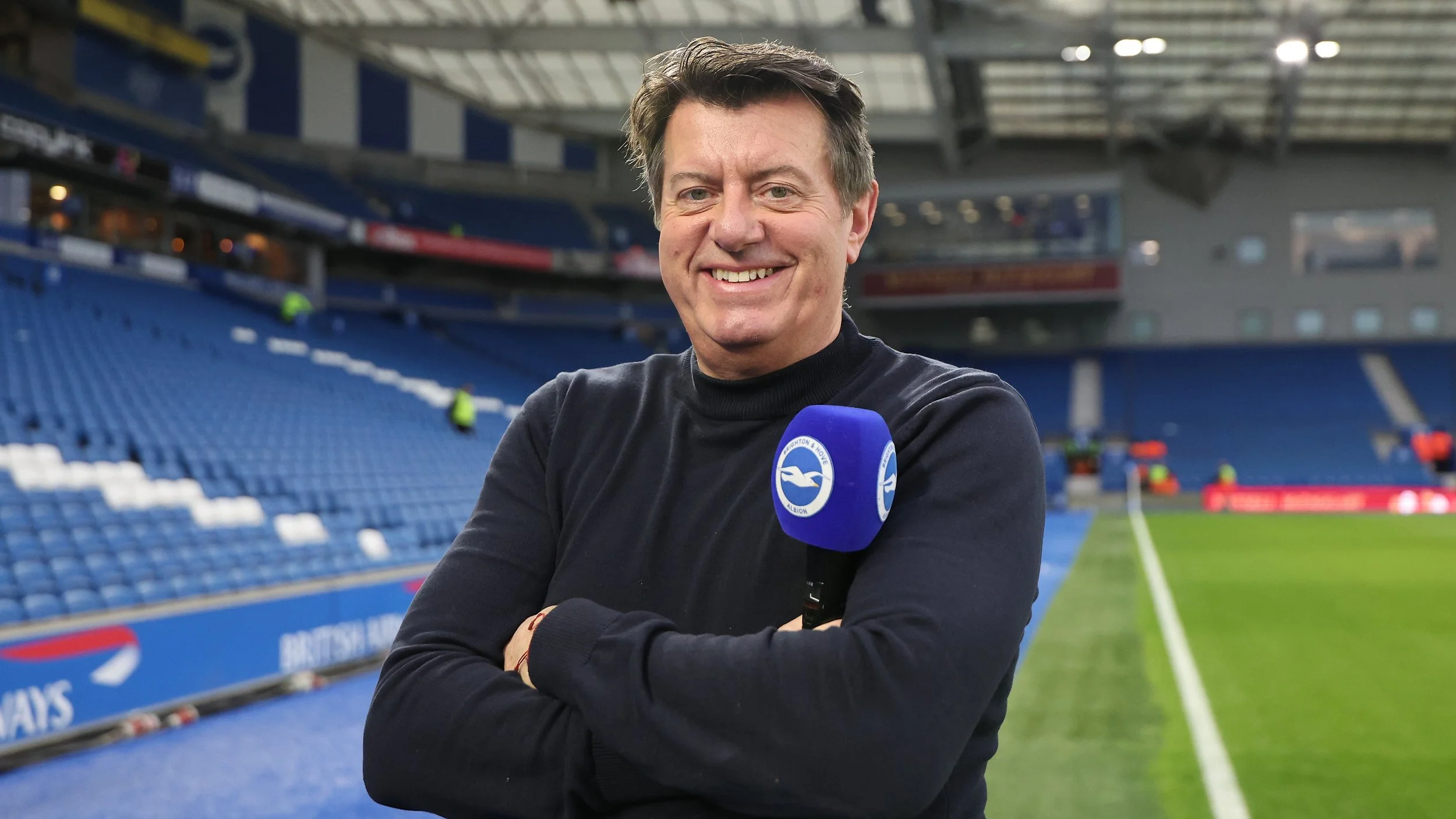 A man holding a Brighton & Hove Albion FC microphone, standing in a football stadium.