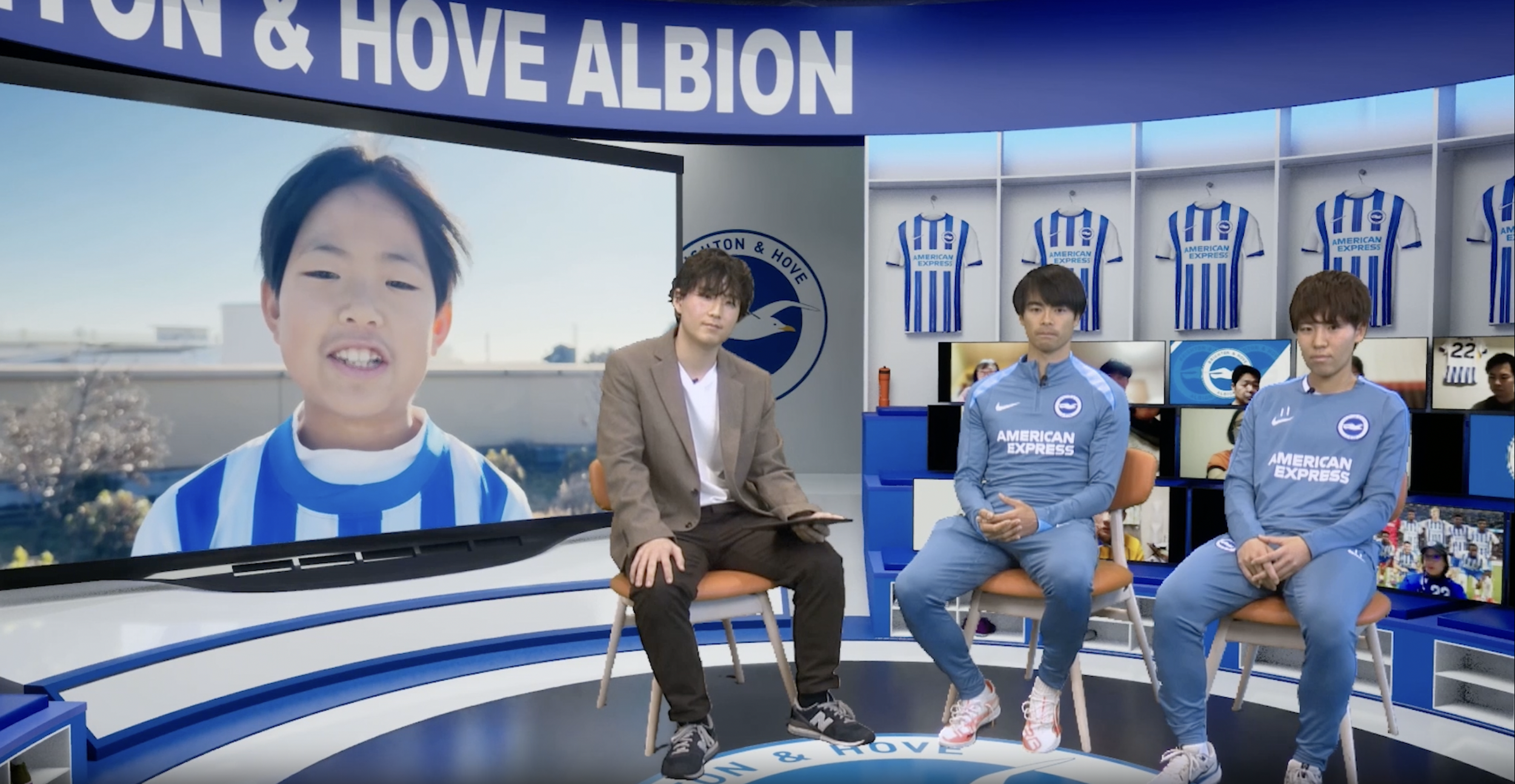 A television studio set featuring two men in blue Brighton & Hove Albion football tracksuits sitting on the right, a woman in a blazer and white shirt sitting on the left, and a large screen behind them showing a child's face in a blue and white striped football jersey, with a Brighton & Hove Albion logo and football jerseys displayed on the walls.