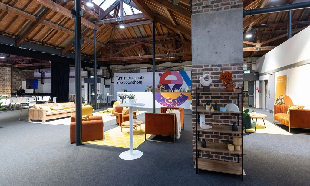 Modern office or event space with high wooden ceiling, brick columns, and contemporary furniture, including armchairs, sofas, and tables, decorated with artwork and sculptures.