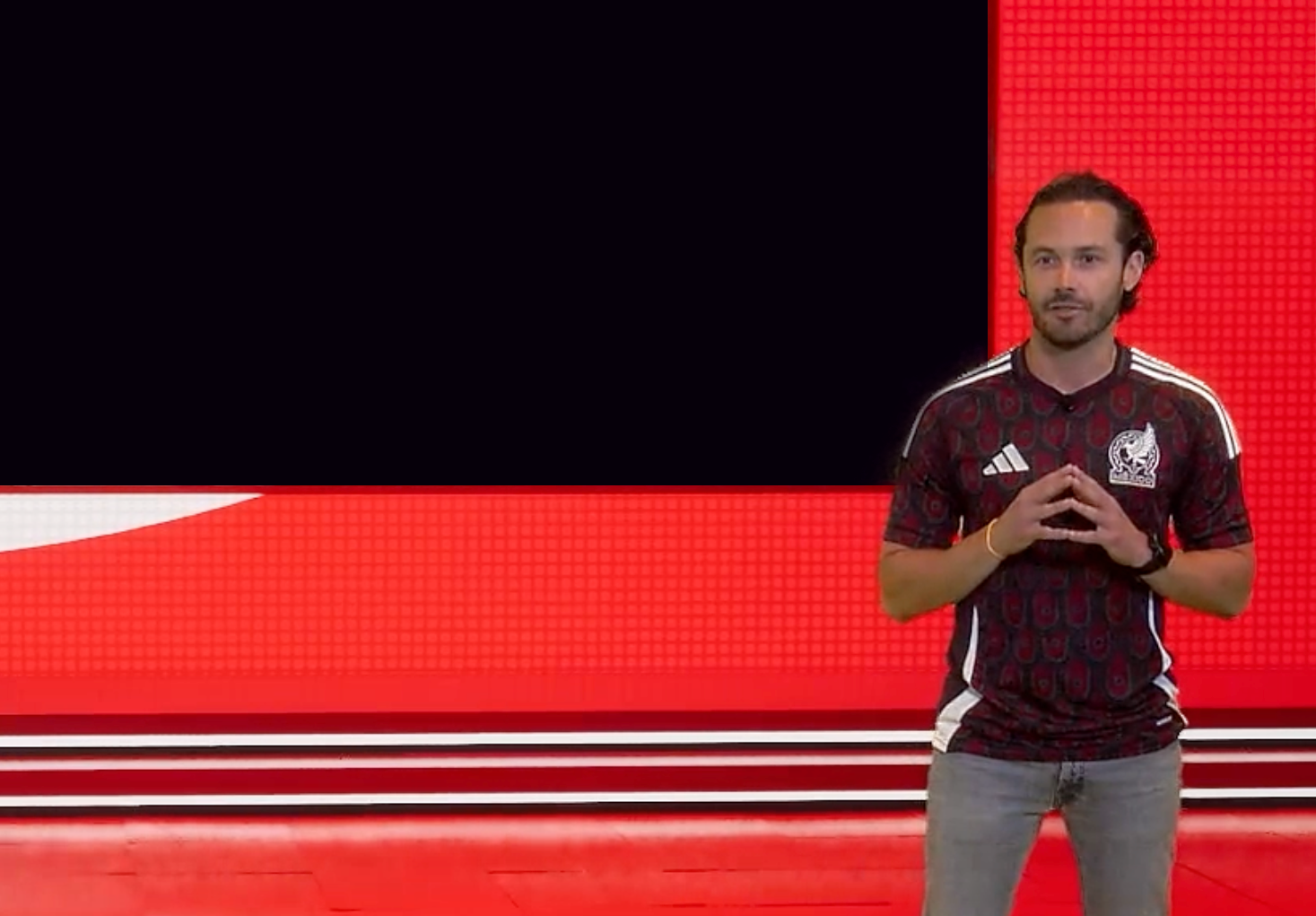 A man standing in front of a large black screen with a red and white background. He is wearing a black sports jersey with white stripes and a logo on the chest, and gray pants. He has dark hair, a beard, and is making a gesture with his hands.