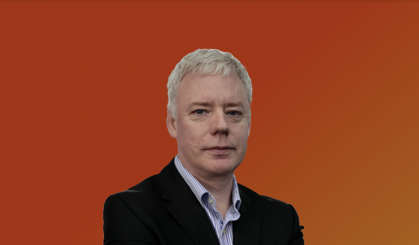 A man with short blonde hair, wearing a black blazer and a blue striped shirt, standing against a red-orange gradient background.