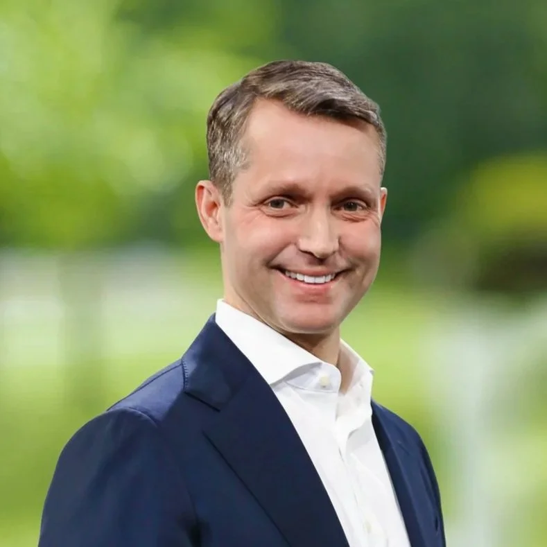 A man with short brown hair wearing a Navy suit jacket and white dress shirt, smiling against a blurred outdoor green background.