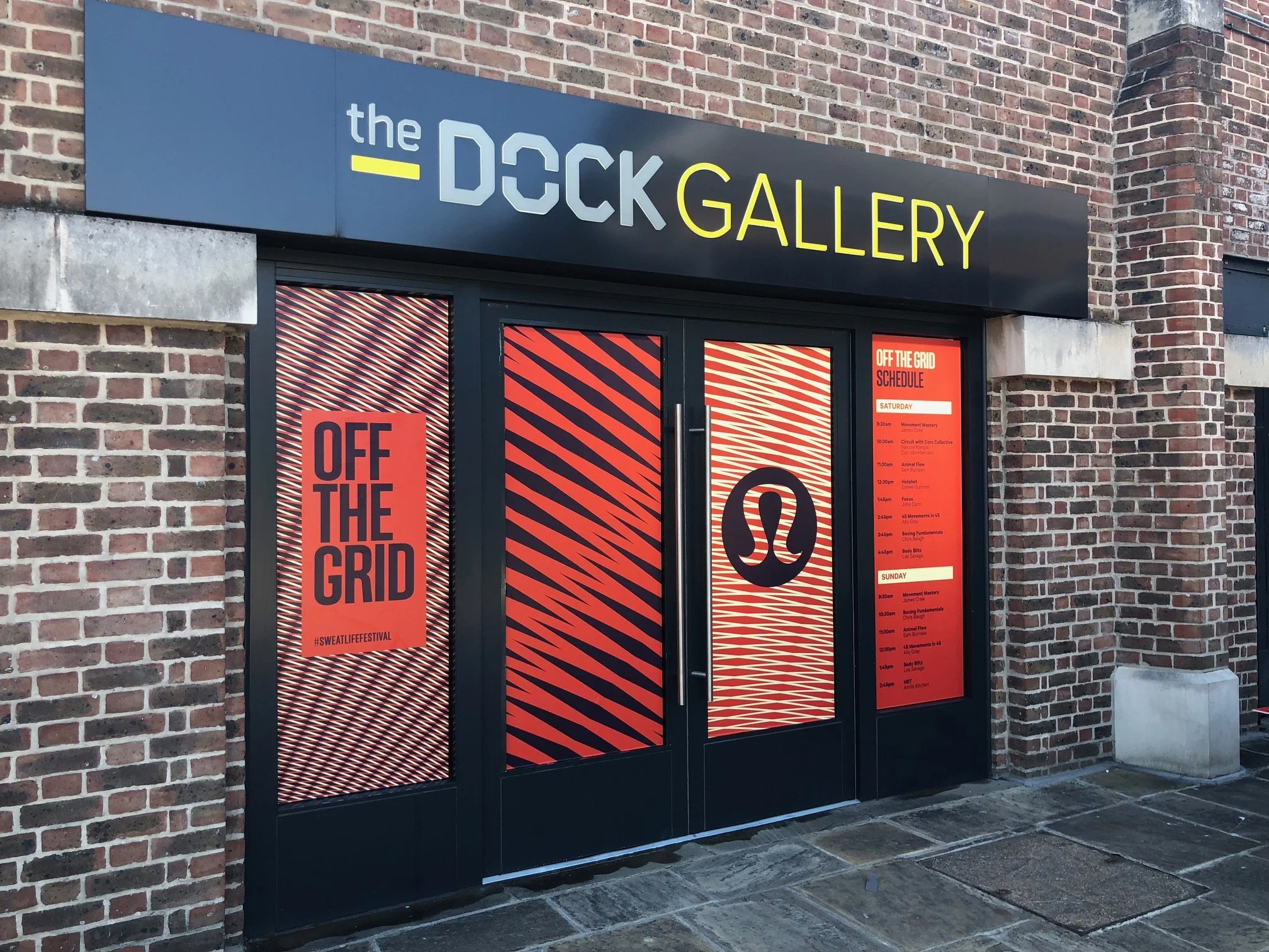 Brick building with a black sign that reads 'the DOCK GALLERY' in large letters, with smaller event schedule and promotional posters on the door.