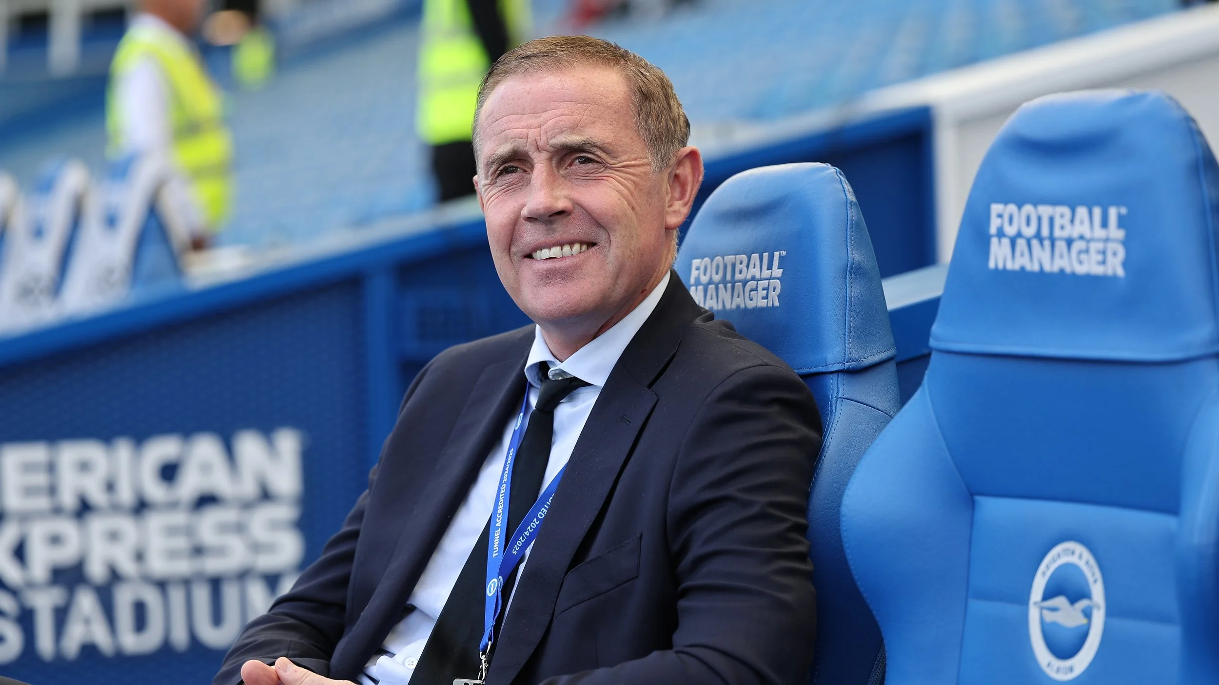 A man in a suit sitting on the bench in a stadium, with blue seats that read 'FOOTBALL MANAGER' behind him, during a football match.