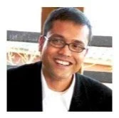 A smiling man wearing glasses, a black blazer, and a white shirt.