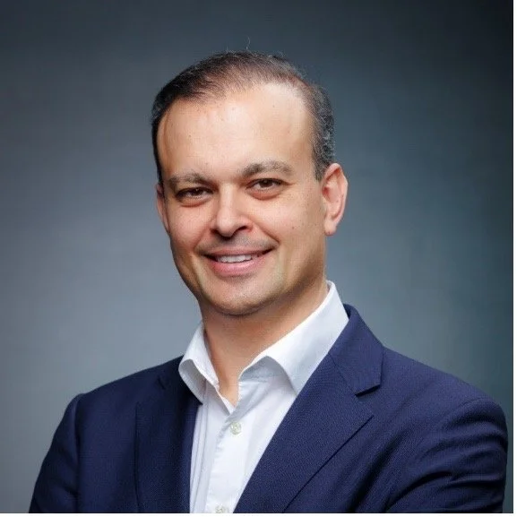 Professional headshot of a man in a dark blue suit and white shirt, smiling, with a gray background.