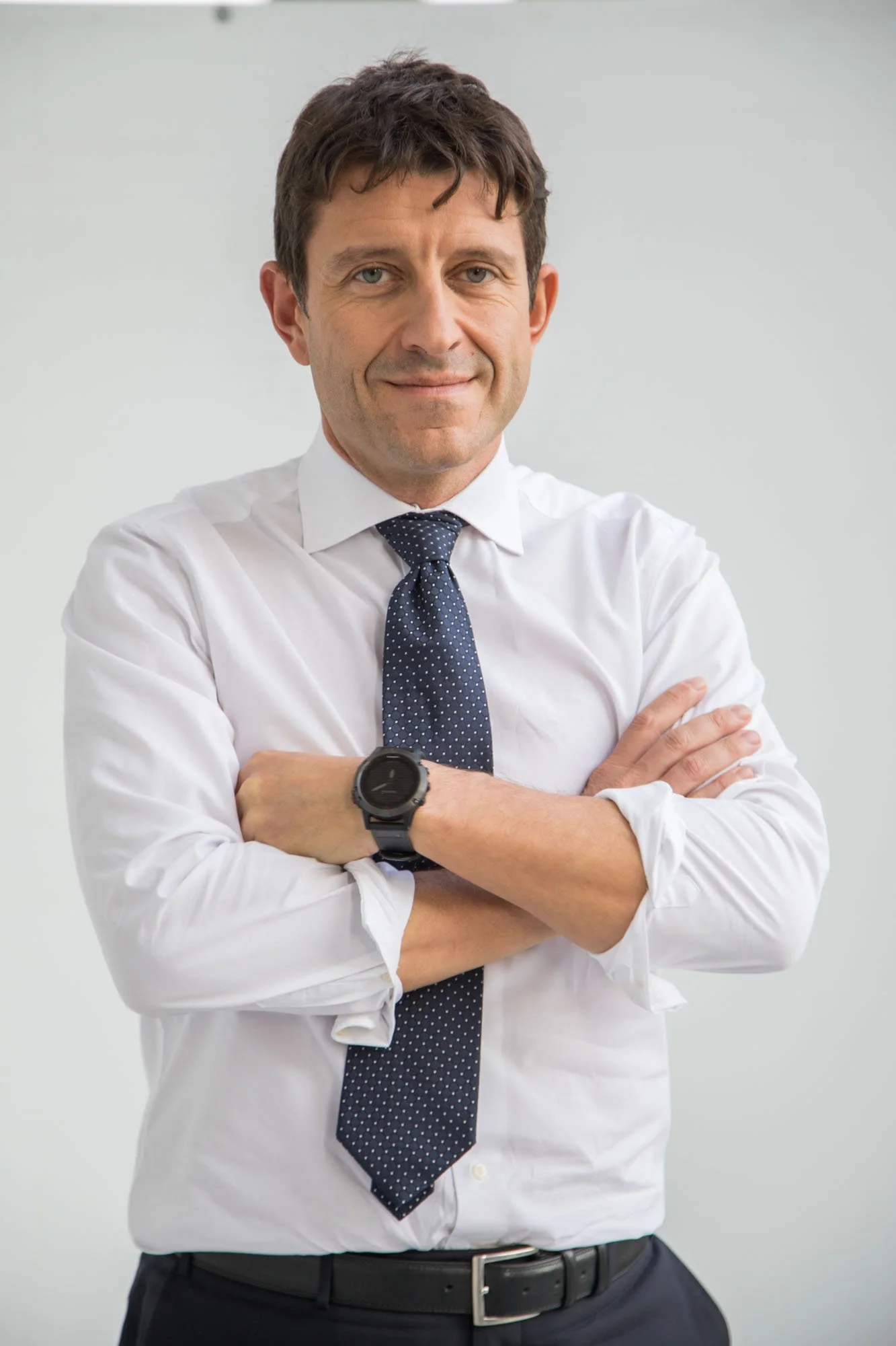 A professional man in a white dress shirt and navy polka dot tie standing with his arms crossed, wearing a black watch on his left wrist, smiling confidently against a plain light gray background.