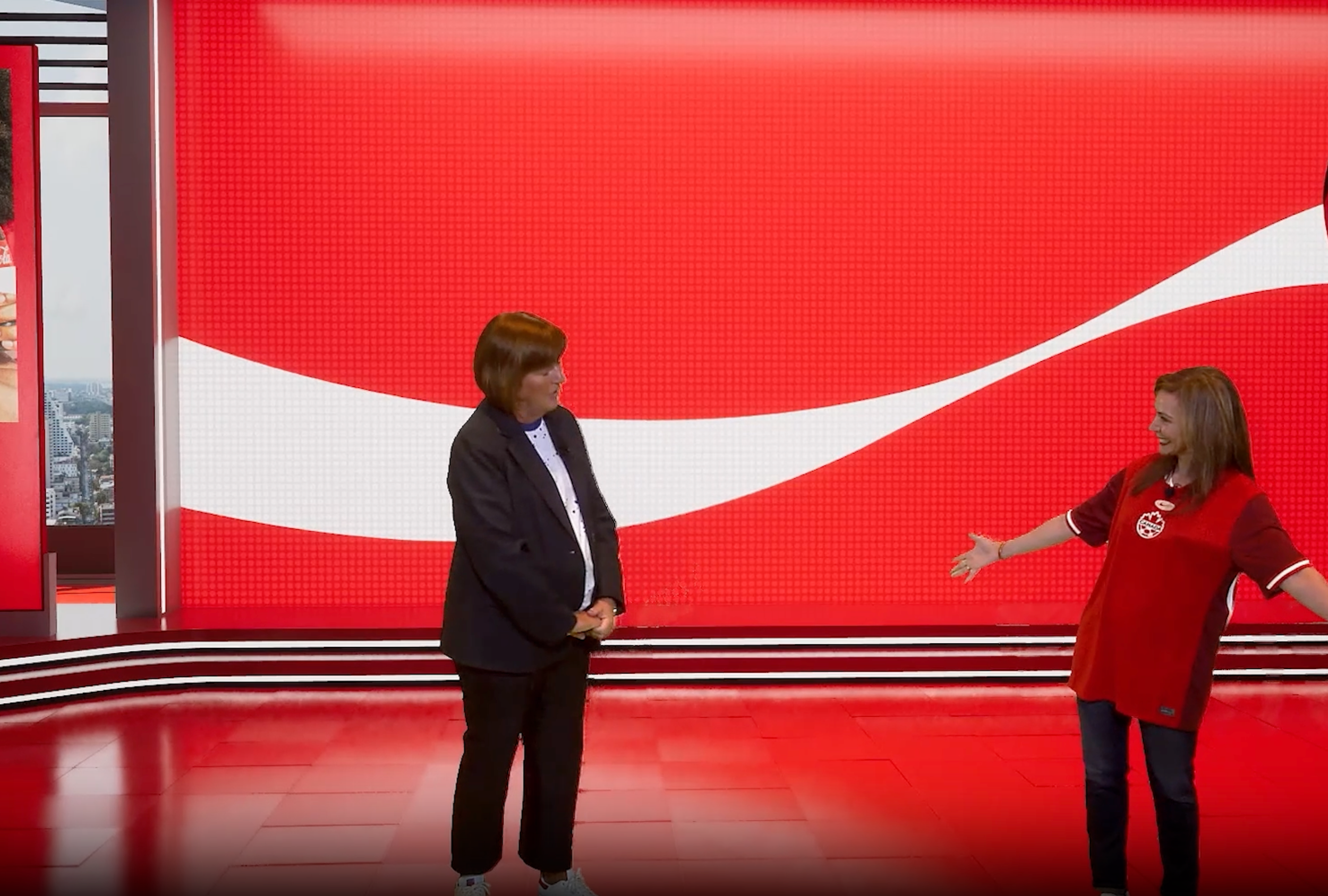Two women standing on a television studio set with a large red and white background design. One woman is dressed in a black blazer and pants, and the other woman is wearing a red sports jersey and jeans, smiling and gesturing with her arms wide open.