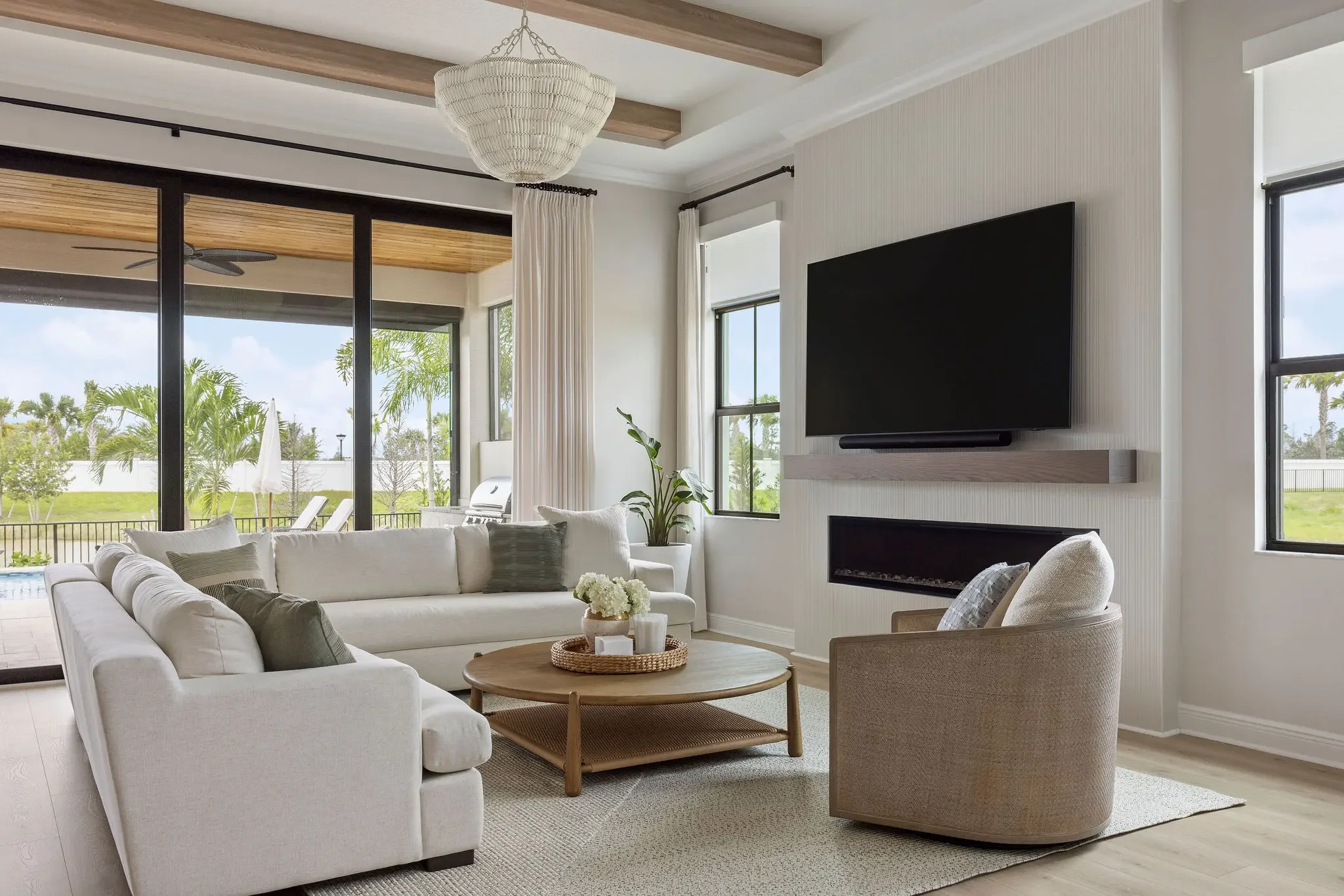 Luxury vacation home living room in Jupiter FL with beaded chandelier, fireplace feature wall, cream sectional, and custom ceiling beams by Interiors by Casa Mia.