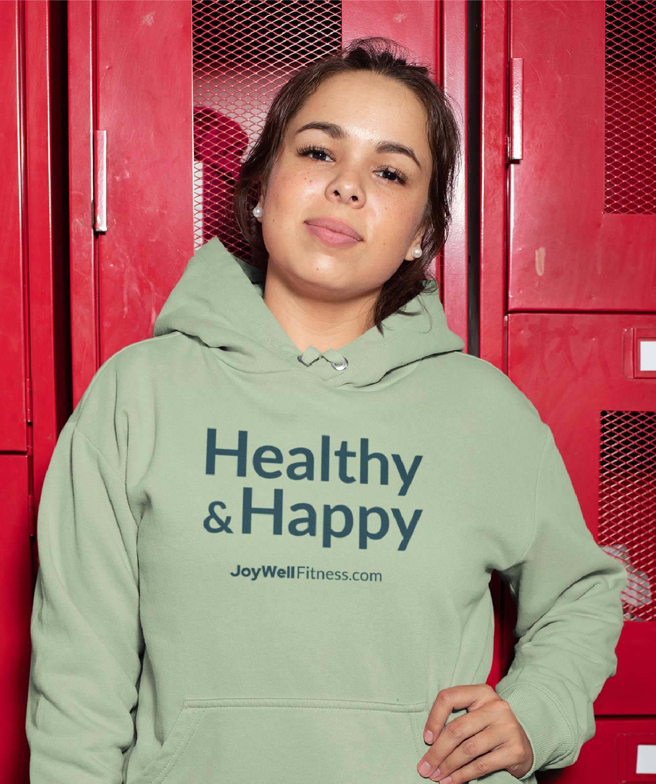 Young woman standing in front of red lockers, wearing a light green hoodie with the words 'Healthy & Happy' and 'JoyWellFitness.com' printed on it, smiling confidently.
