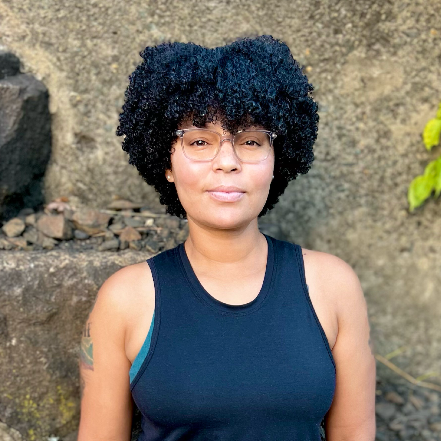 JoyWell Fitness Founder, Mi'Kele Rodriguez, A woman with glasses and curly black hair, wearing a black sleeveless top, standing outdoors against a rocky background with some green leaves visible.