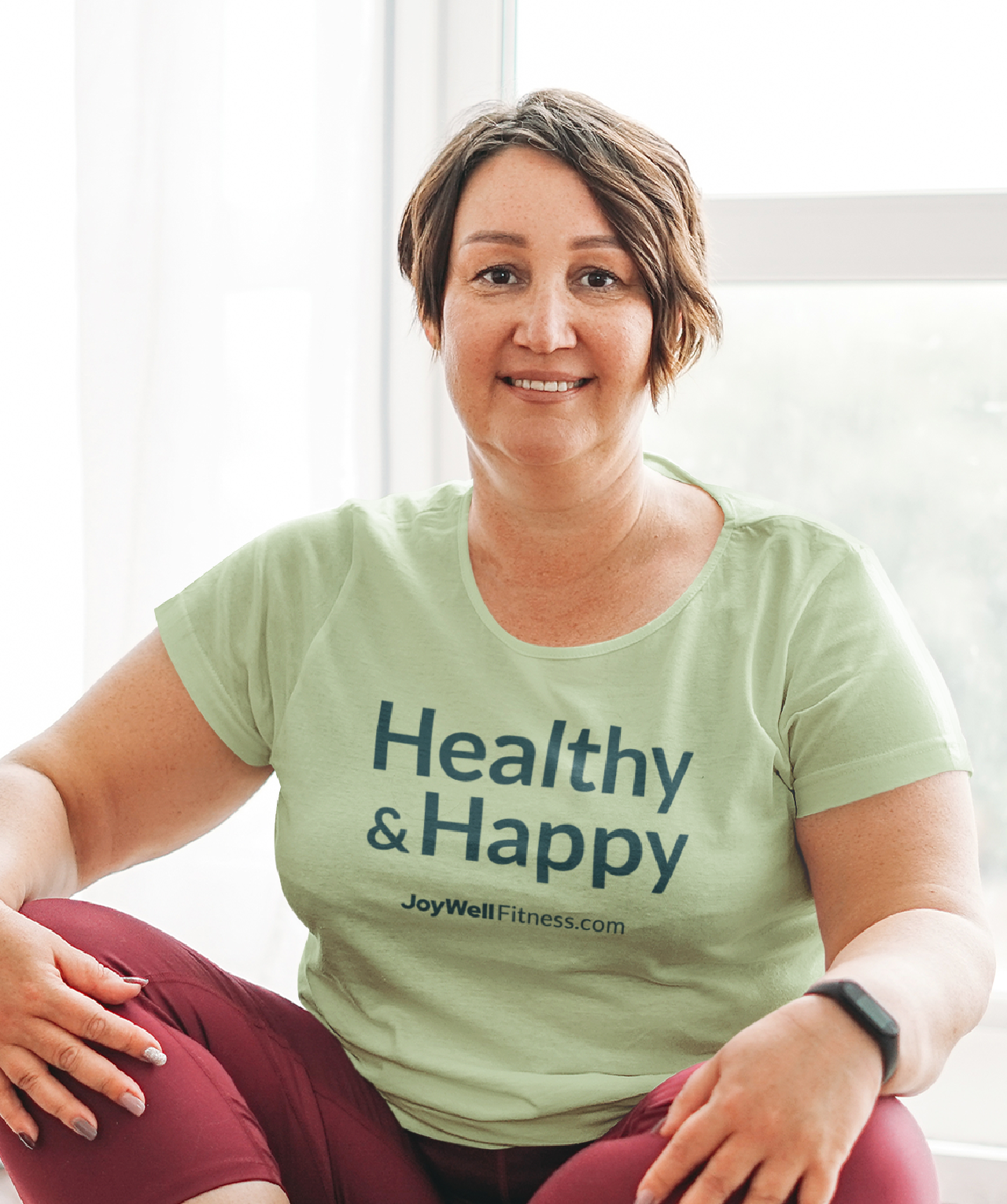 A woman sitting cross-legged indoors near a window, smiling, wearing a light green JoyWell Fitness t-shirt with the text 'Healthy & Happy' and maroon pants.