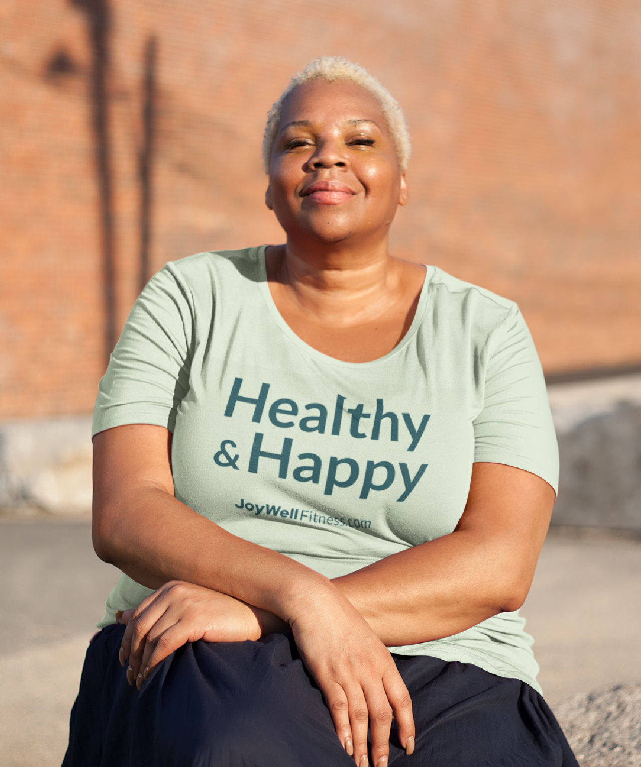A woman with short, blond hair smiling outdoors. She is wearing a light green JoyWell Fitness  T-shirt that says 'Healthy & Happy' and has her arms crossed, sitting on a rock with a brick wall in the background.