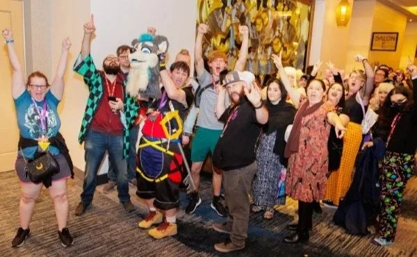 Group of people at a convention, some raising hands with peace signs, posing with a costumed mascot, all indoors near a gold decor background.