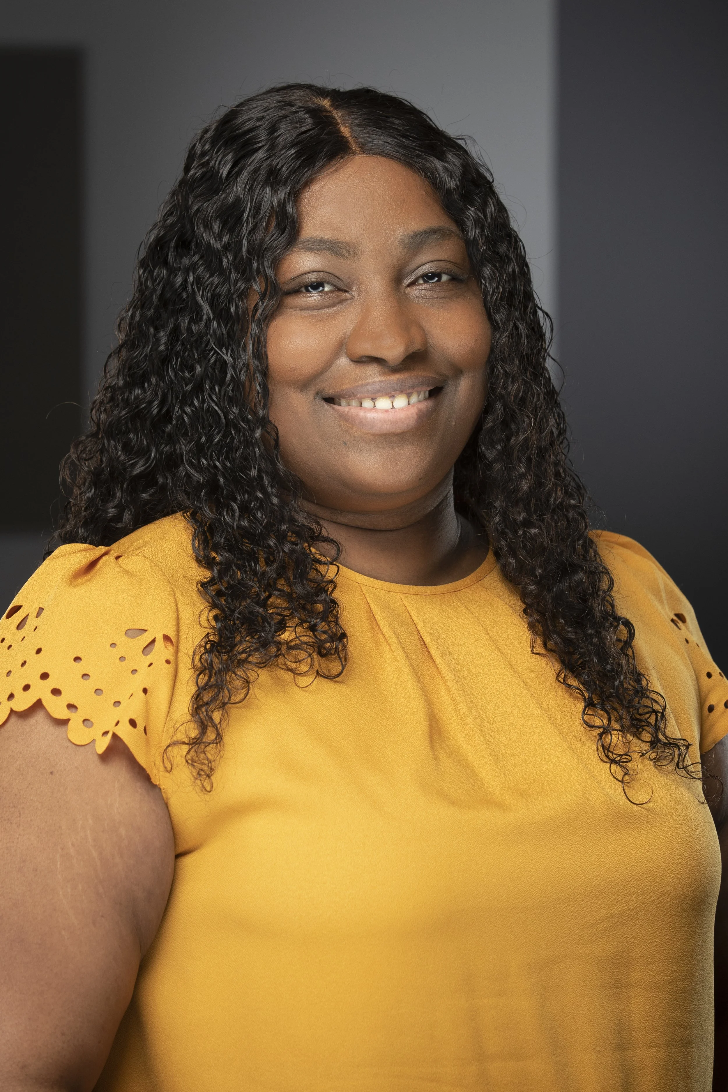A woman with dark, curly hair wearing a yellow top with scalloped sleeve details, smiling at the camera against a dark background.
