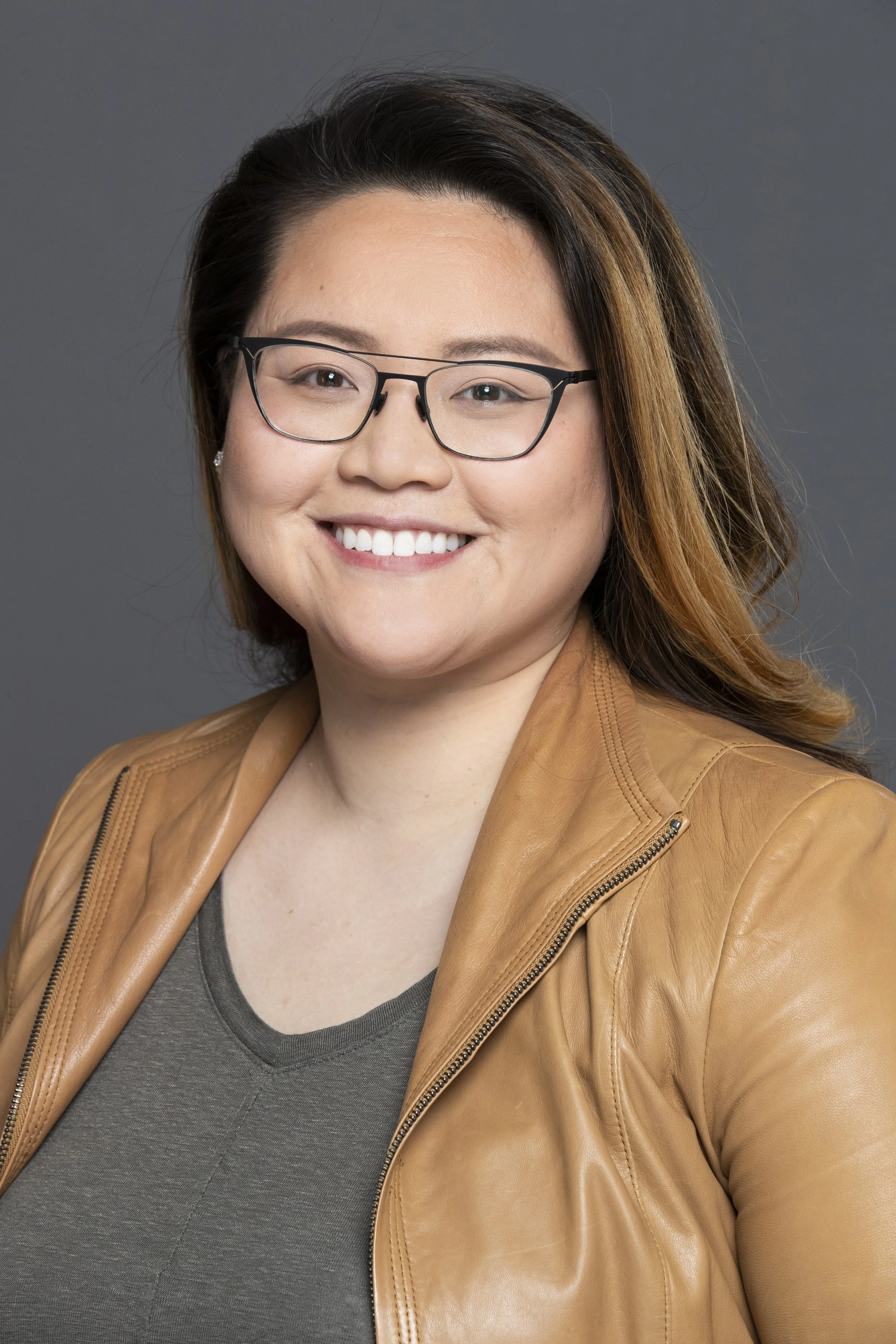 A woman with glasses smiling, wearing a tan leather jacket and a grey top, against a grey background.