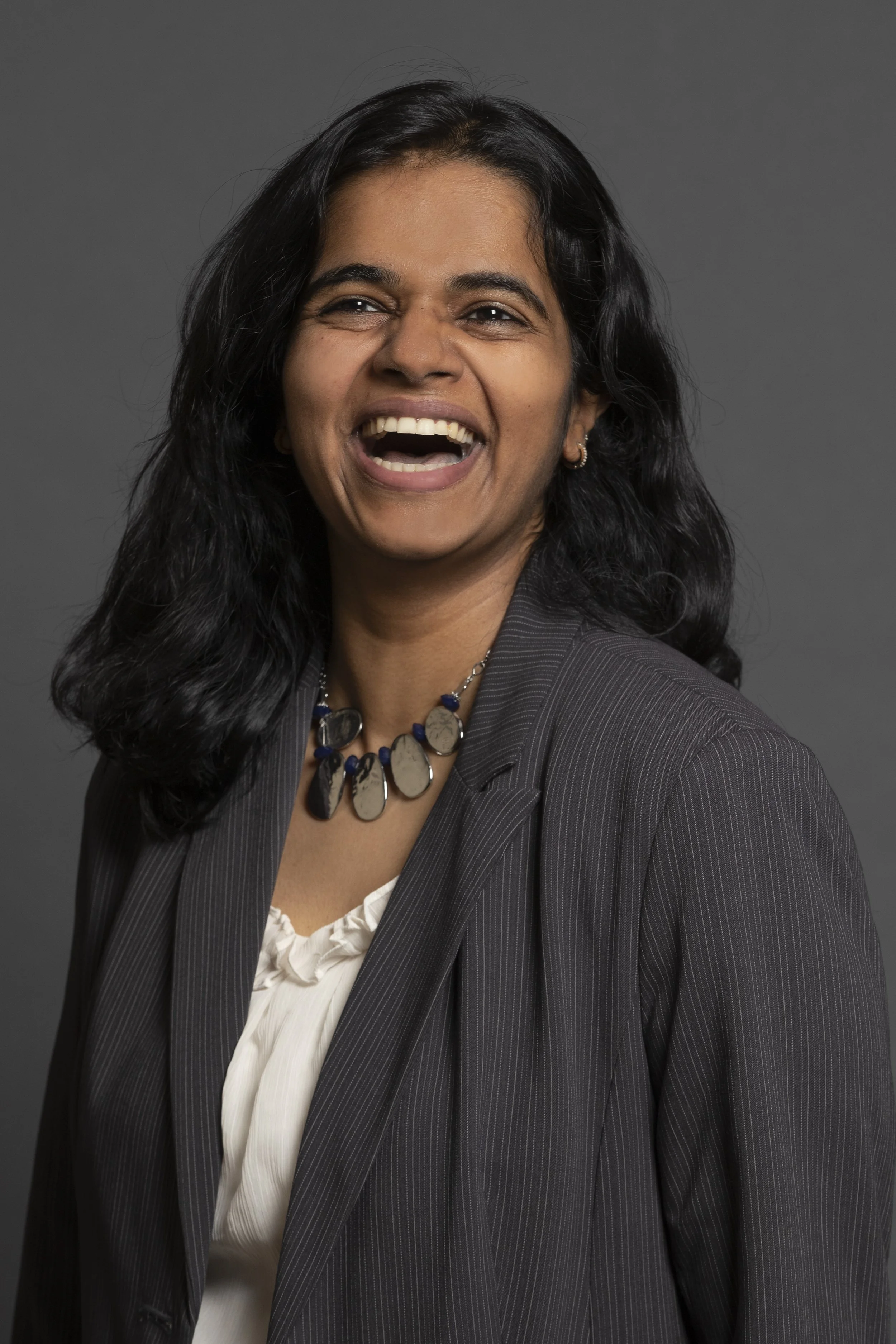 A woman with long black wavy hair wearing a dark pinstripe suit, white blouse, and a beaded necklace, smiling and laughing against a gray background.
