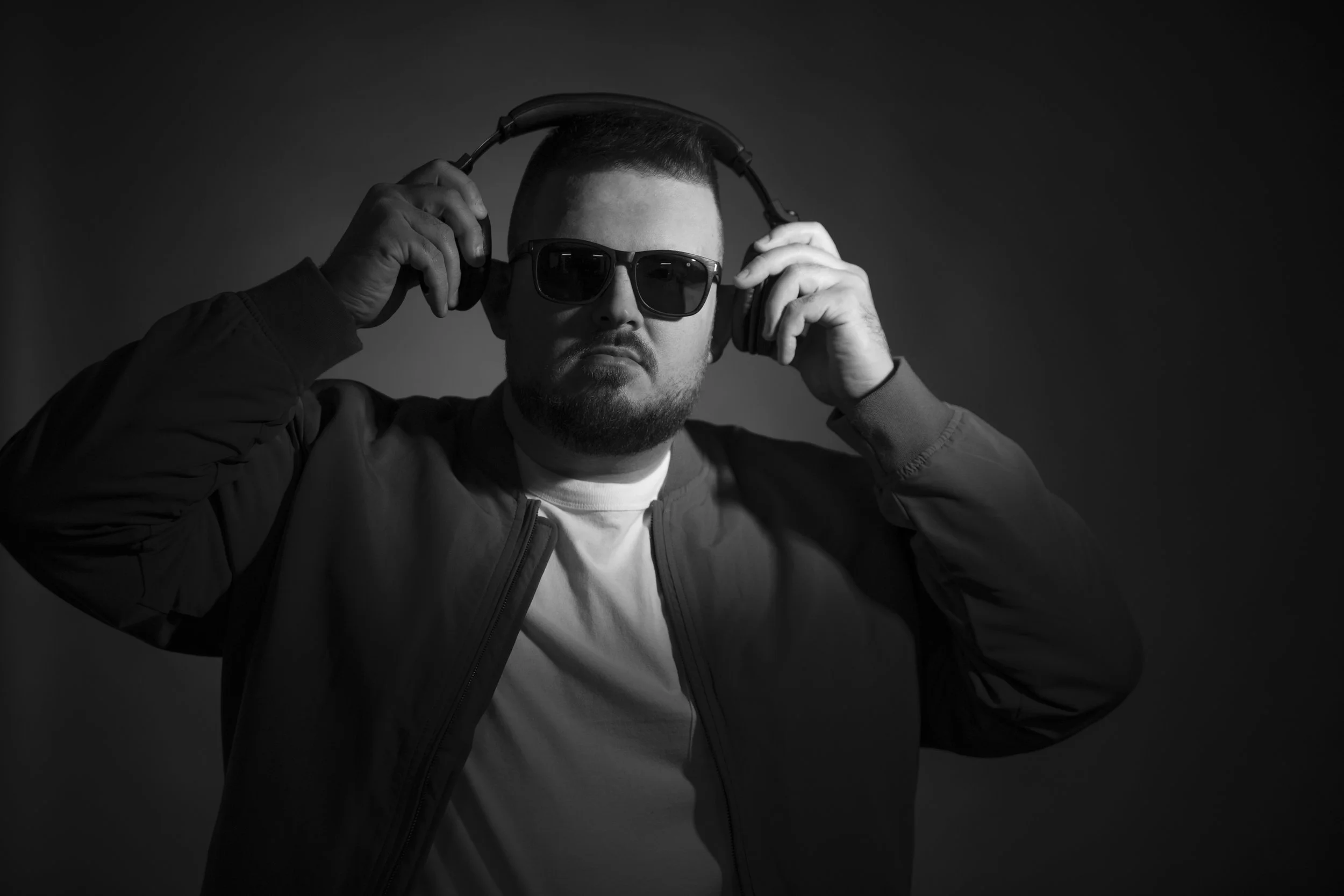 A man wearing sunglasses, a casual jacket, and a T-shirt, holding headphones to his ears in a black and white photo.