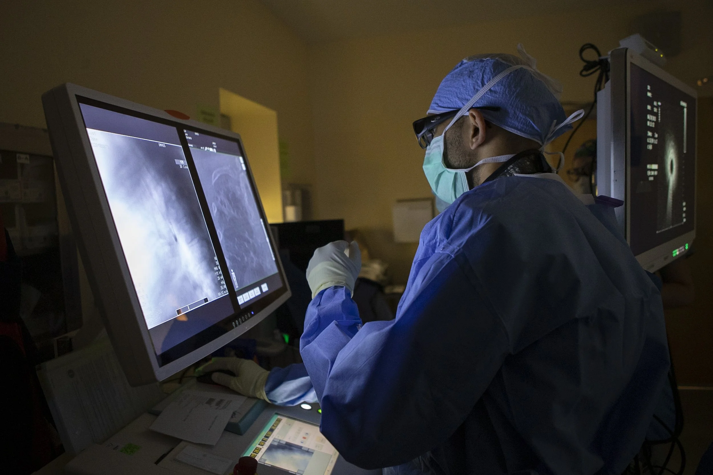 A medical professional in scrubs, mask, and protective gear examining X-ray images on computer screens in a dimly lit hospital room.