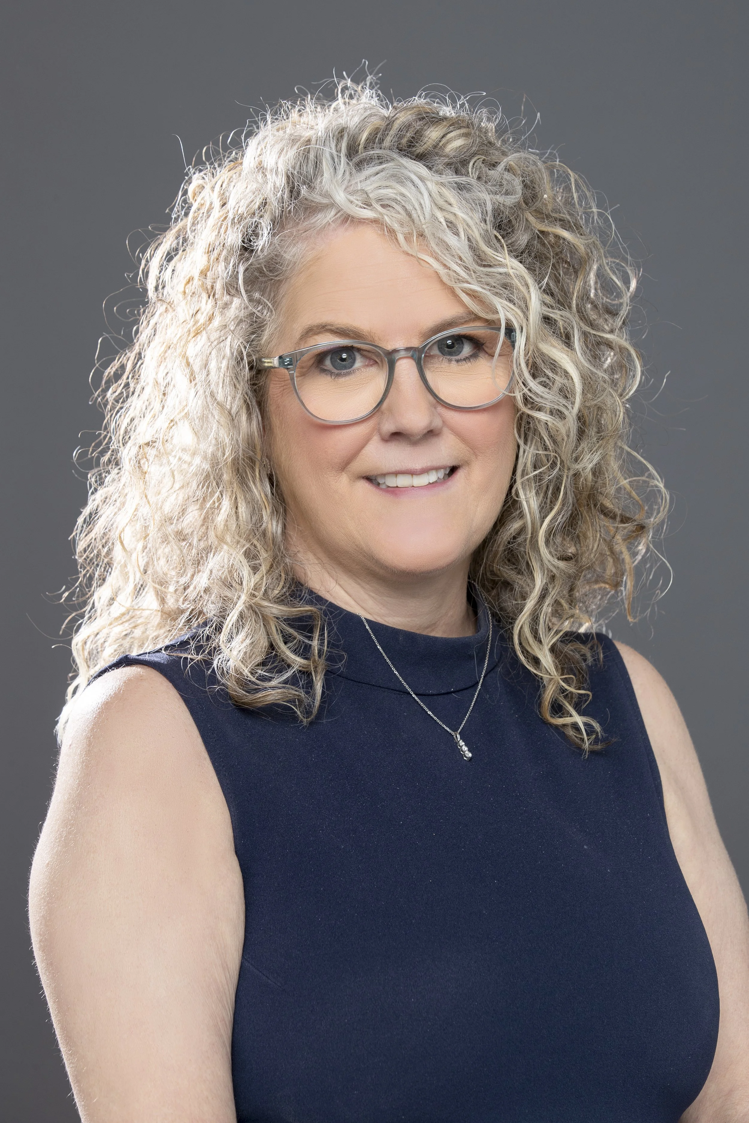 Portrait of a woman with curly blonde hair, glasses, wearing a sleeveless navy blue top, and a silver necklace, against a gray background.