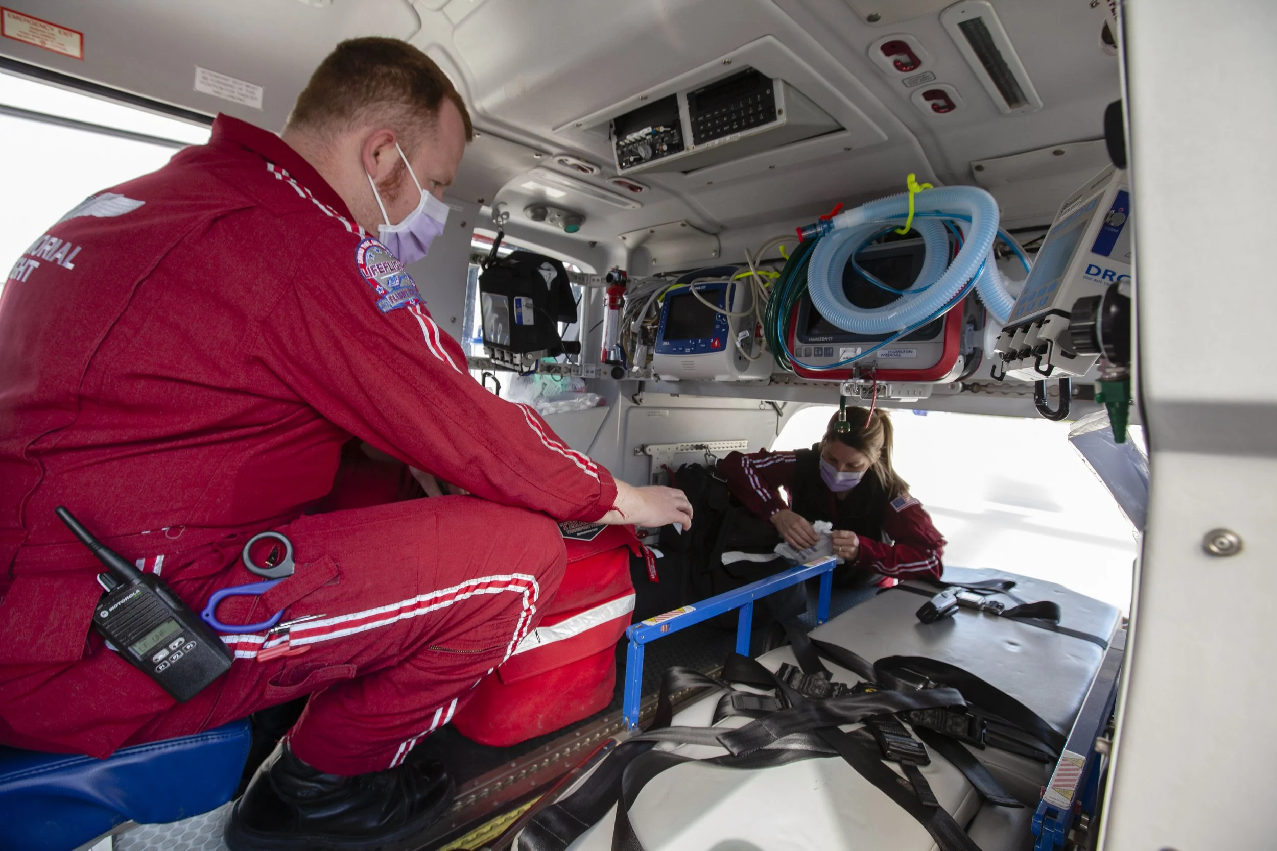 Two emergency medical personnel inside a helicopter, both wearing masks and red uniforms, attending to a patient lying on a stretcher.