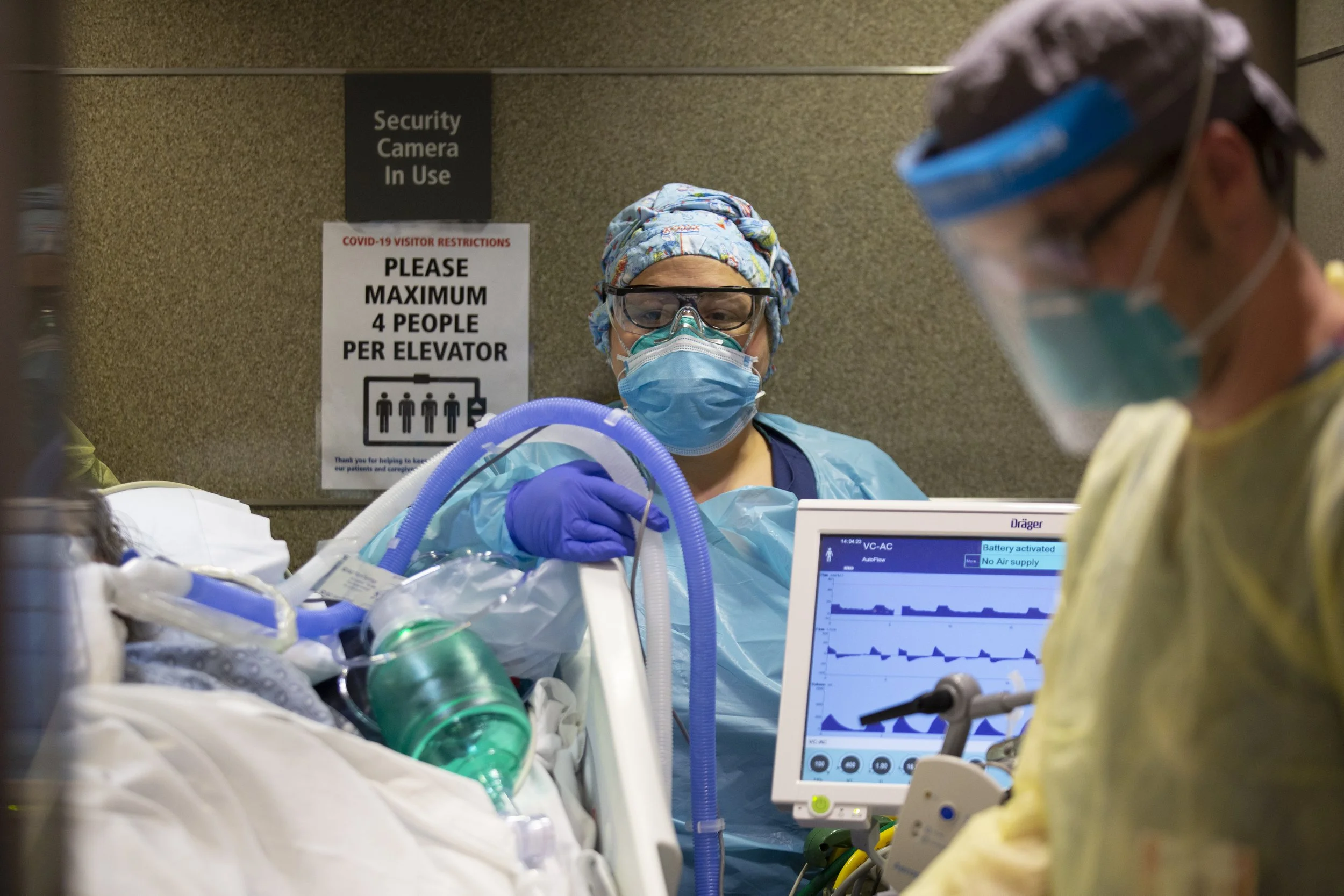 Medical healthcare workers wearing masks, gloves, and protective gear attending to a patient in a hospital emergency room. One worker is operating a monitor showing vital signs.