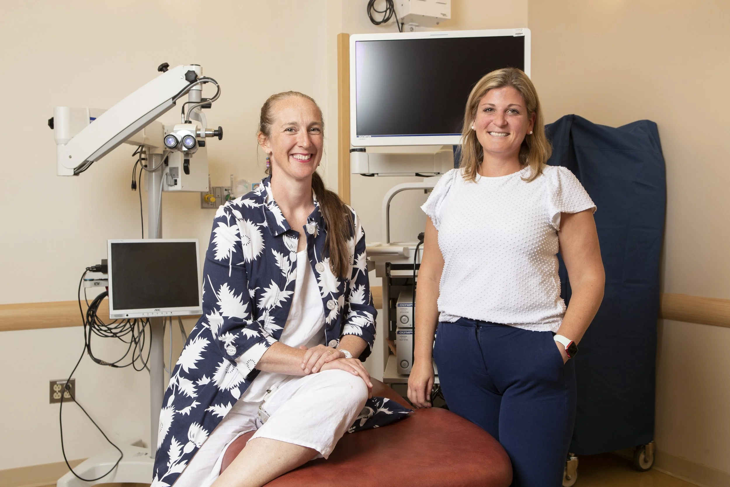 Two women in a medical examination room, one seated on an exam table and the other standing, smiling, with medical equipment and monitors in the background.