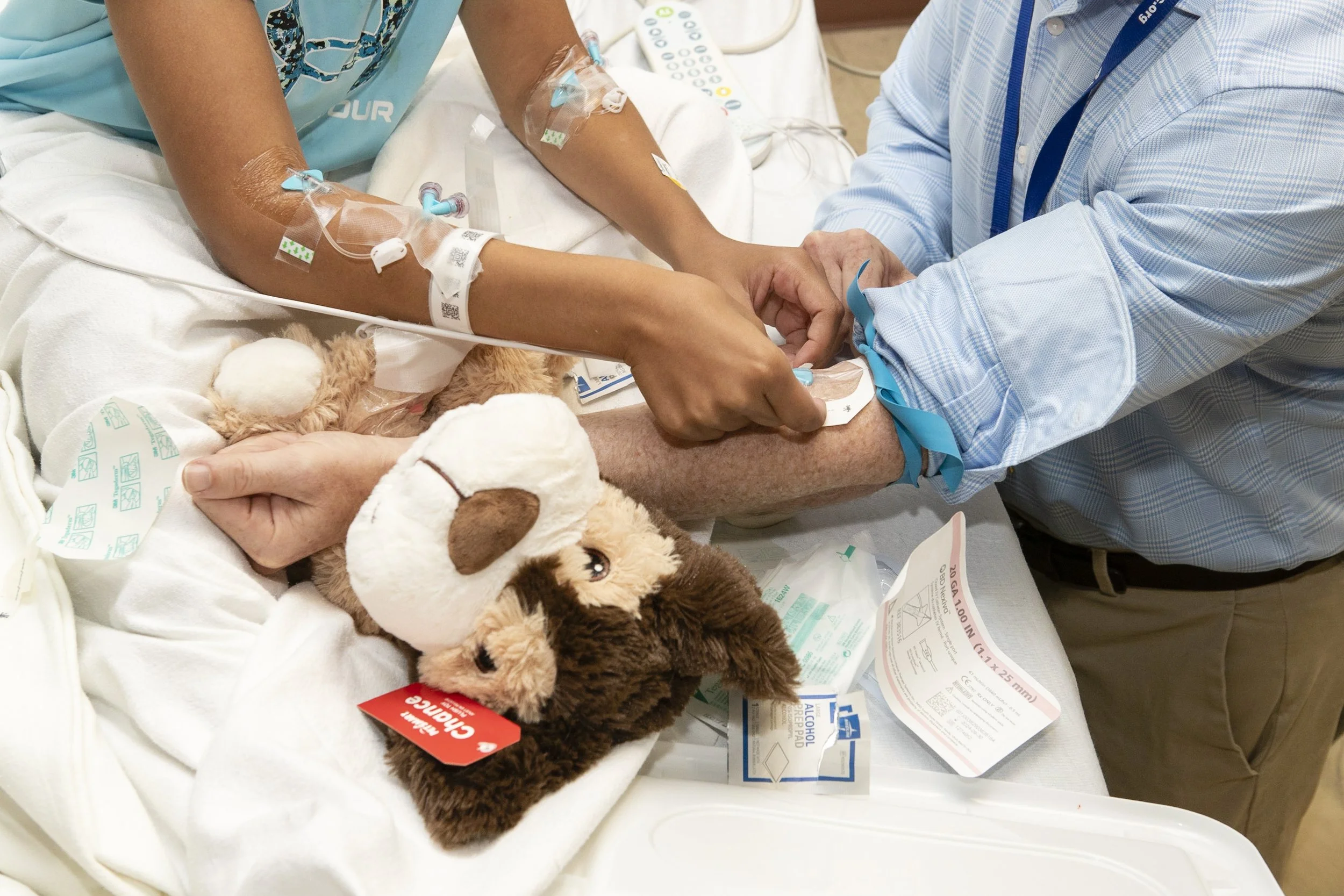 A person in a hospital bed receiving an intravenous injection from another individual, with a stuffed dog toy placed on the bed.