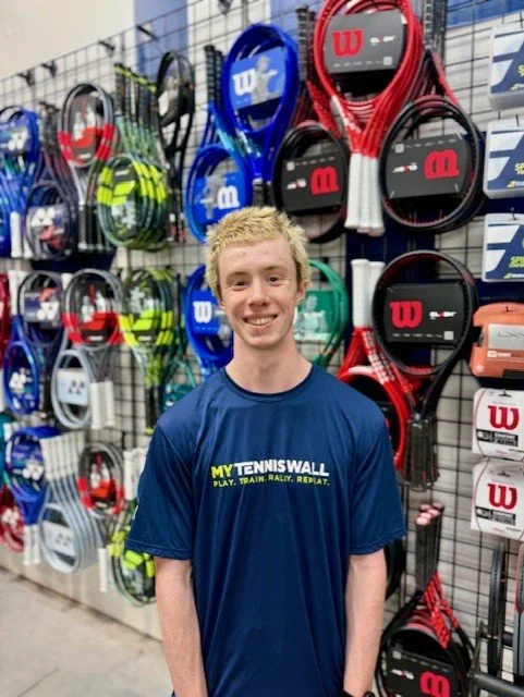 Young man with blonde hair smiling and wearing a blue T-shirt in a tennis store, with tennis rackets displayed on the wall behind him.