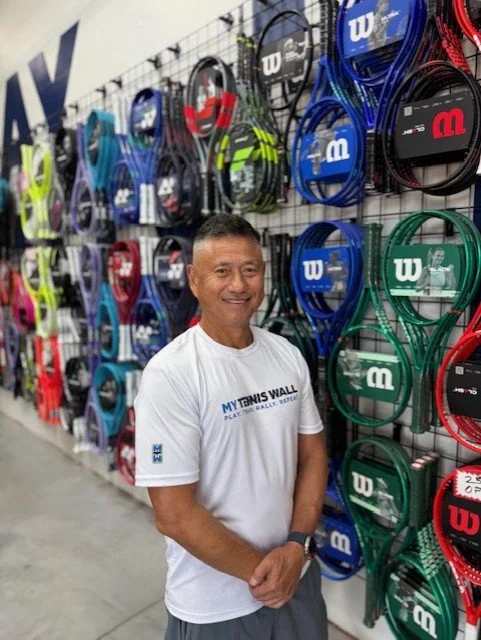 A man in a white T-shirt stands in front of a display of tennis rackets and accessories at a sports store, smiling at the camera.