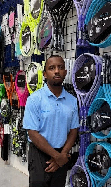 A man in a light blue polo shirt standing in front of tennis rackets hanging on display at an athletic store.