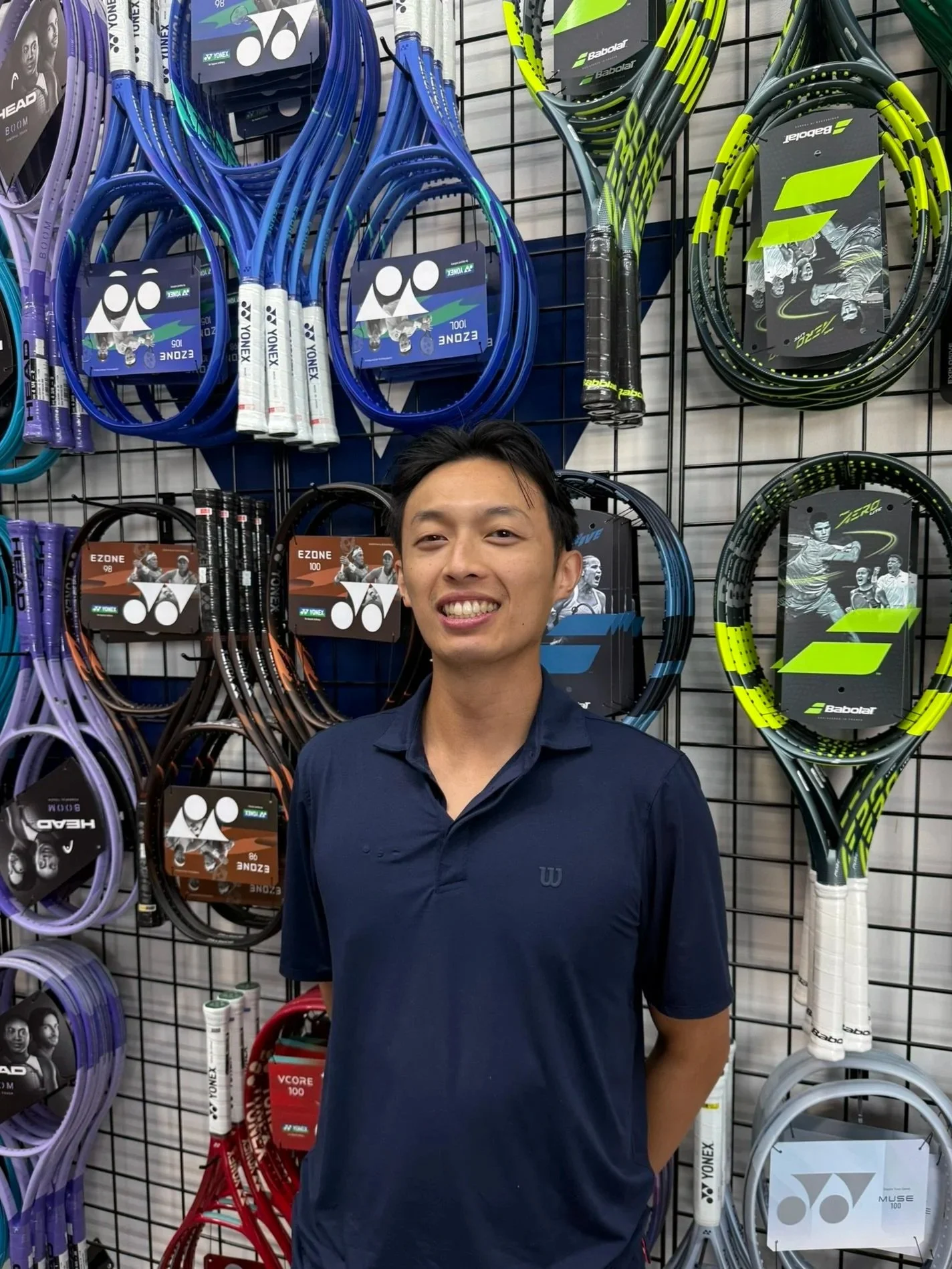A smiling man standing in front of a display of tennis rackets in a sporting goods store.