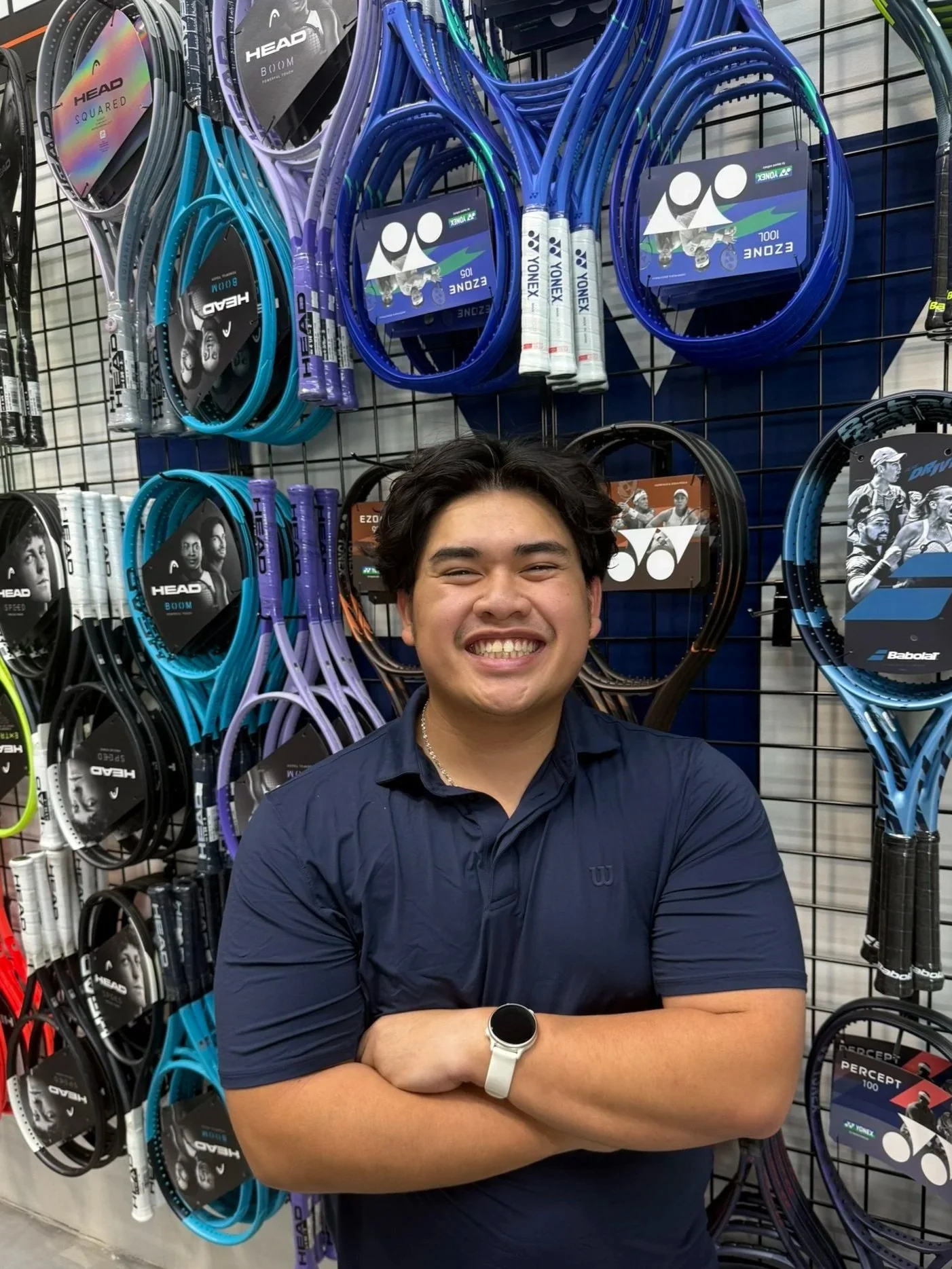 A young man with dark hair smiling, standing in front of a display of tennis racquets at a sporting goods store.