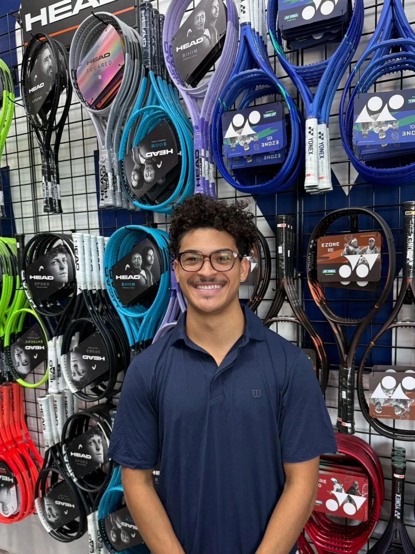 Smiling young man with glasses and curly hair standing in front of a display of tennis rackets at a sports store.