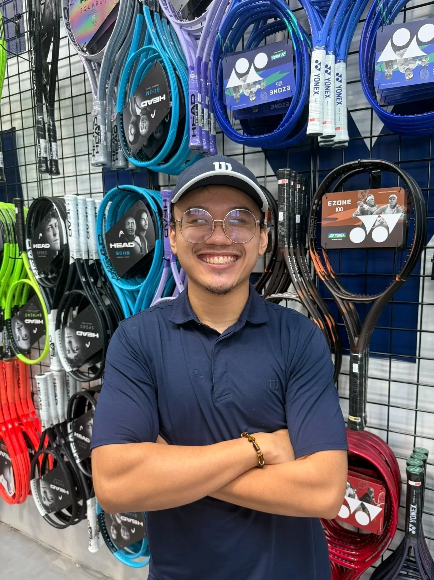A smiling man with glasses and a black cap, standing with arms crossed in front of a display of various tennis rackets in a sporting goods store.