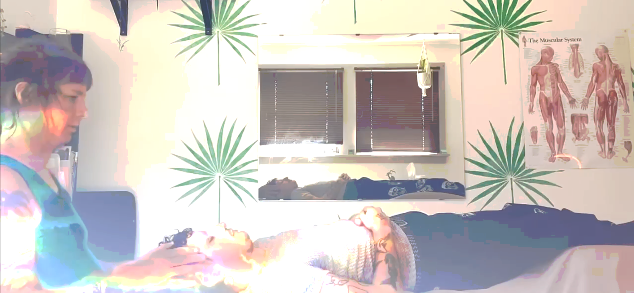 A white woman with hands on the head of a white male bodywork client. The background is sparkly with a mirror and palms decorating the wall. There is a muscle chart on the wall as well.