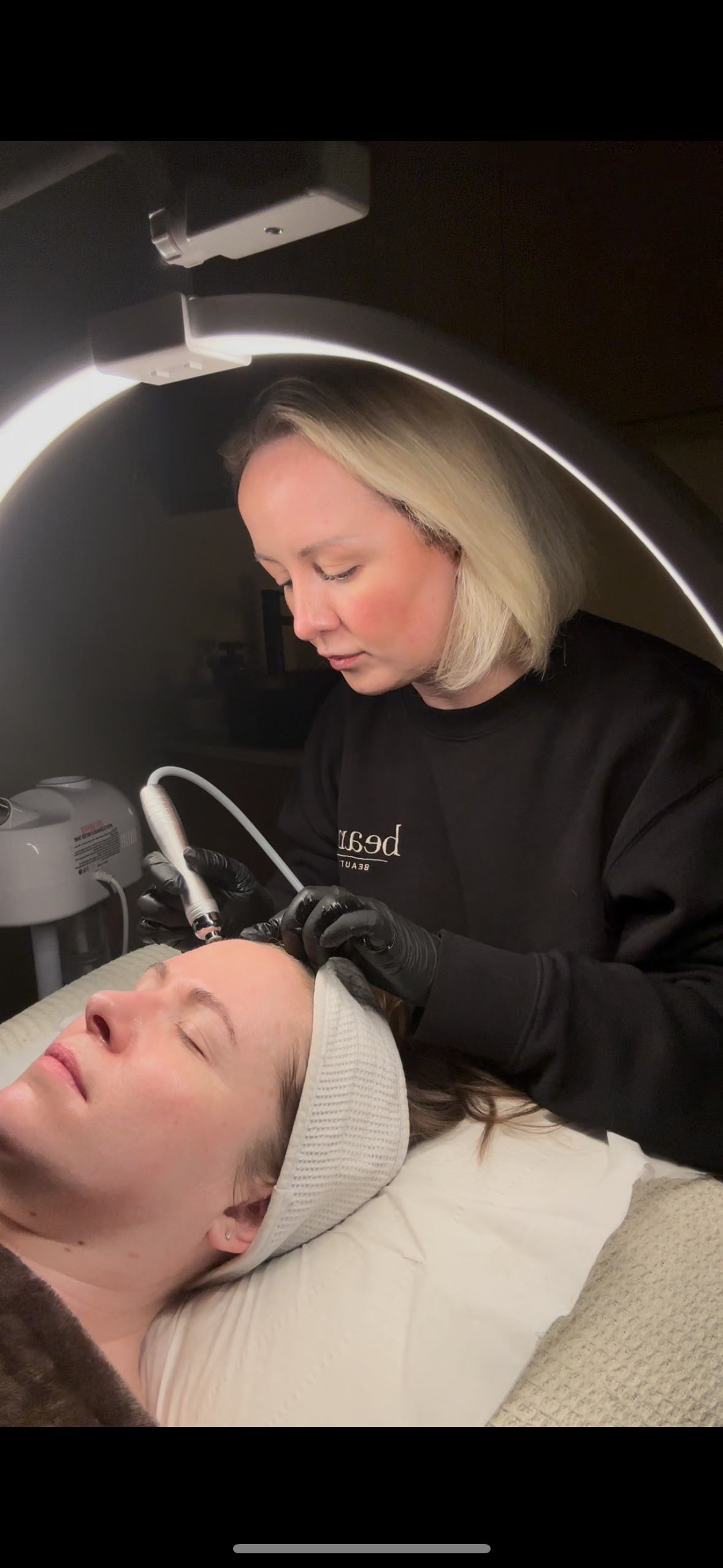 A woman getting a facial treatment or skin procedure by a professional under a specialized light or laser device in a skincare clinic.