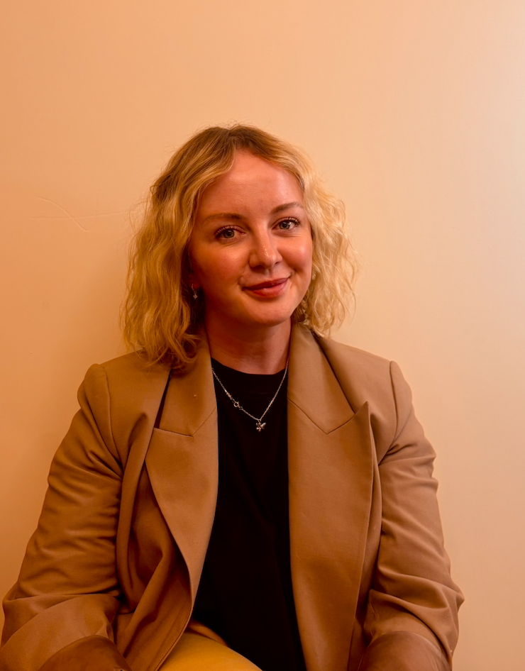 A woman with shoulder-length blonde hair wearing a beige blazer and black top, sitting against a plain beige wall.
