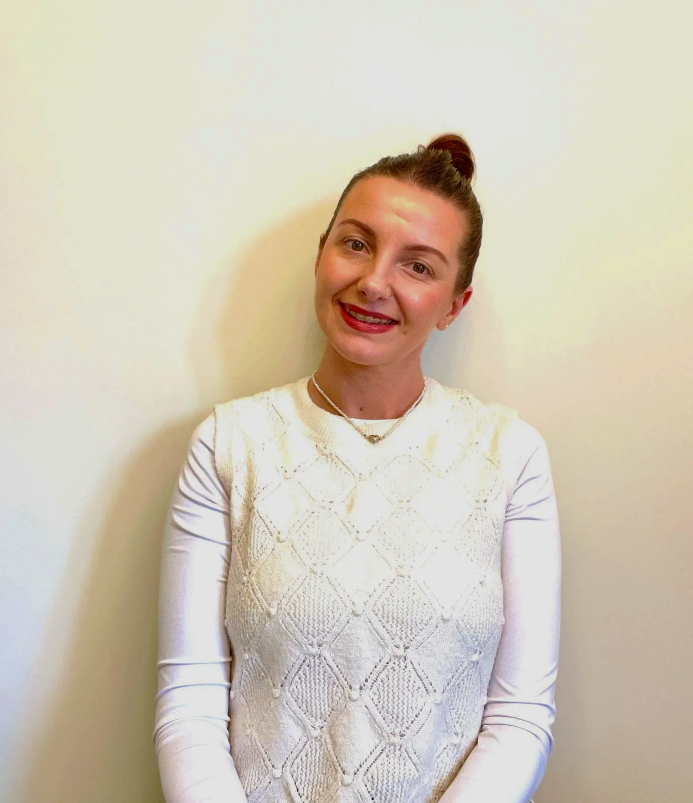 A woman smiling at the camera, standing against a plain light-colored wall, wearing a white long-sleeve top with a diamond pattern, a necklace, and her hair tied up in a bun.