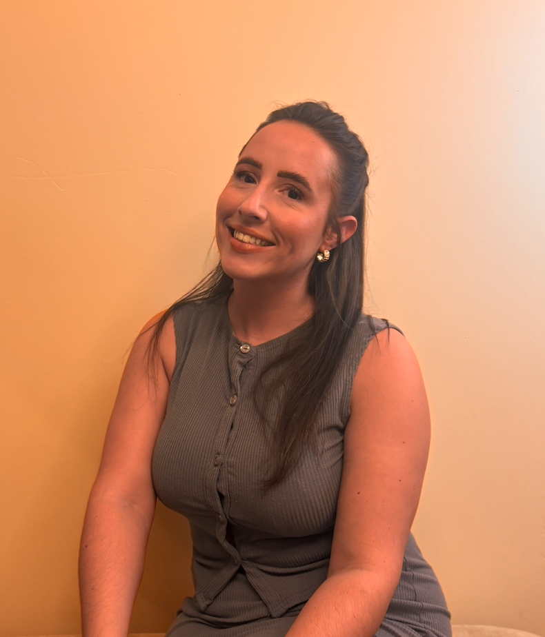 A woman with long dark hair, smiling, wearing a sleeveless gray top, sitting against a beige wall.