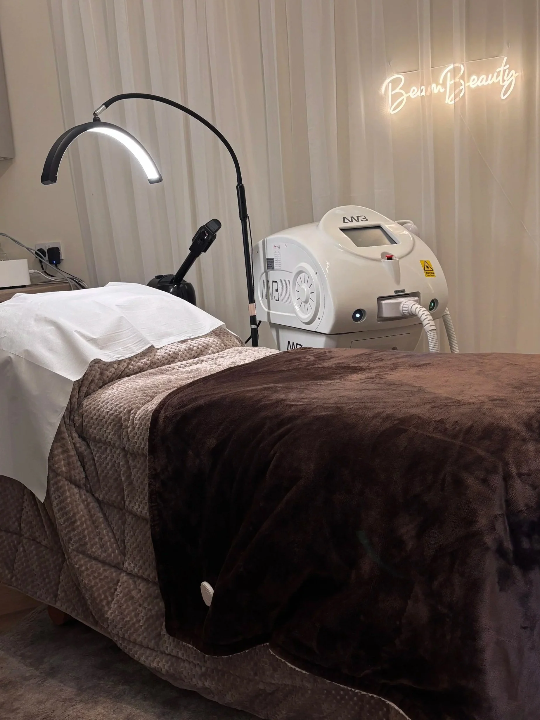 A spa treatment room with a bed covered in brown and beige blankets, a face cradle with a white pillow, and a medical device with tubes and a digital display on the side. In the background, a wall with a neon sign that reads 'Be au tify' and light-colored vertical paneling.