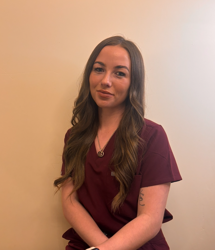 A young woman with long wavy brown hair, wearing maroon scrubs and a necklace, posing against a plain beige wall.