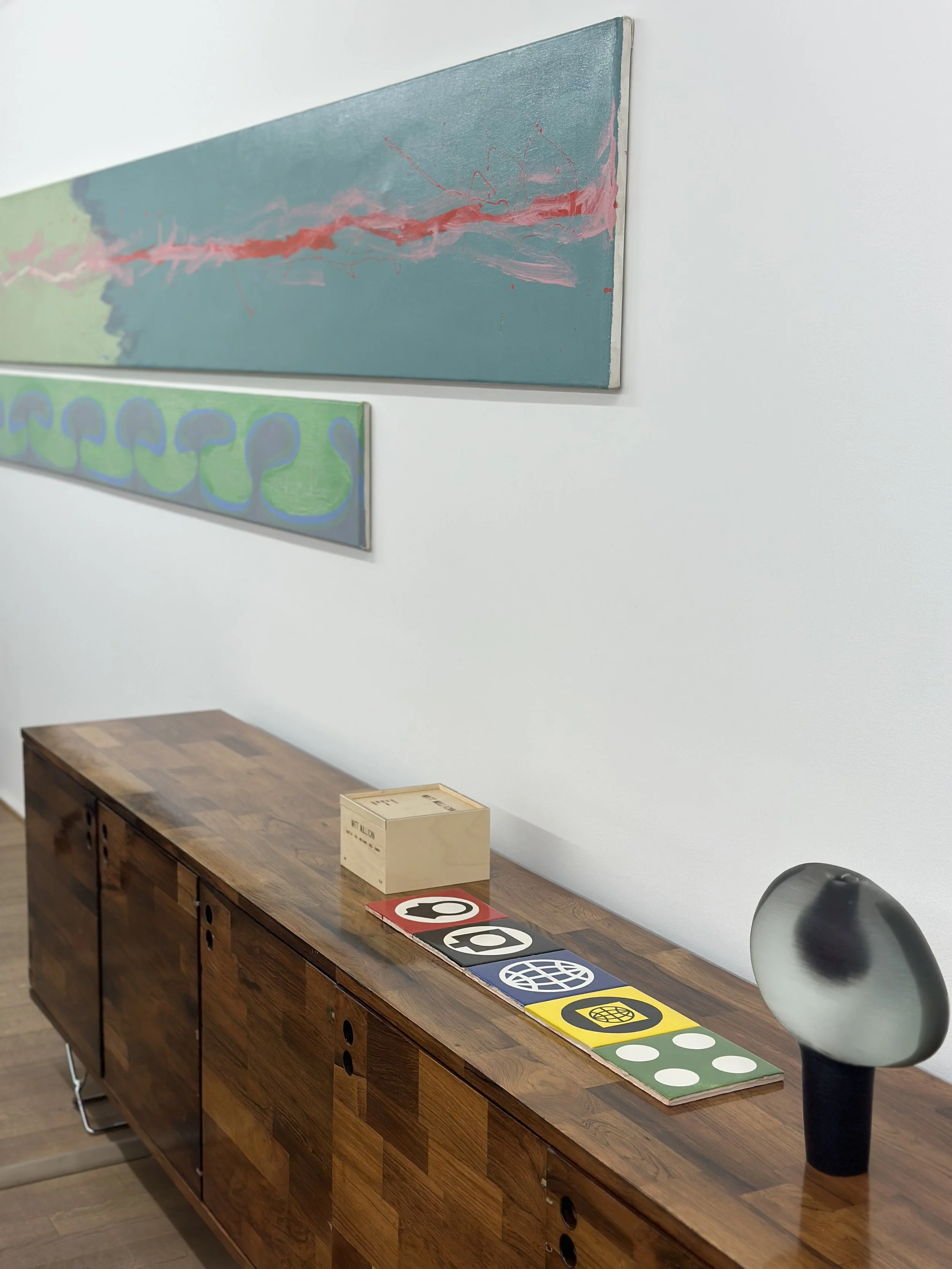 An art gallery interior with abstract paintings on the wall, a wooden sideboard with graphic design magazines or portfolios, a wooden box, and a modern black and white ceramic vase.