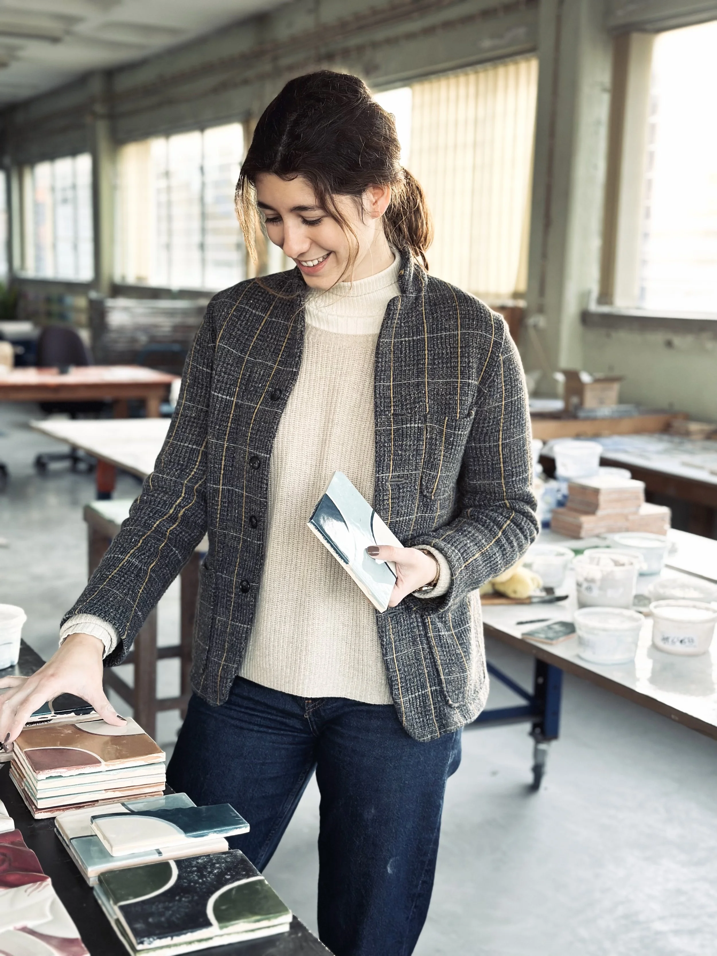 A woman in a gray plaid jacket and cream turtleneck examining color samples at a workshop or studio with tables of design or fabric materials in the background.