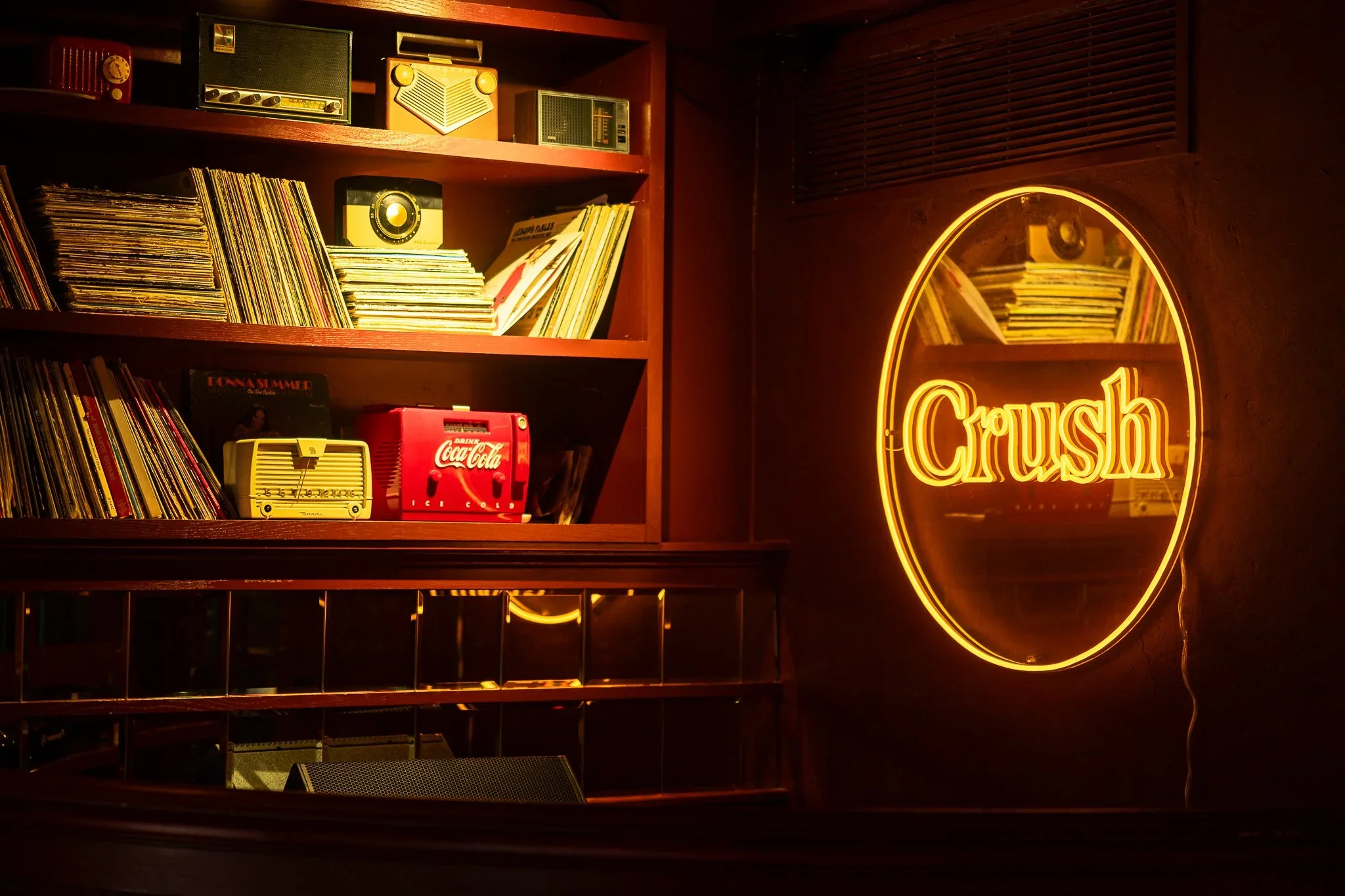 A young man with curly hair and a beard taking a selfie with his phone in a music lounge or bar. He is wearing a black t-shirt that says 'Crush Dance Bar,' black shorts, and a watch. The background features a colorful wall decorated with vintage concert posters, a mirror, and some lighting fixtures. There is DJ equipment on a table in front of him.