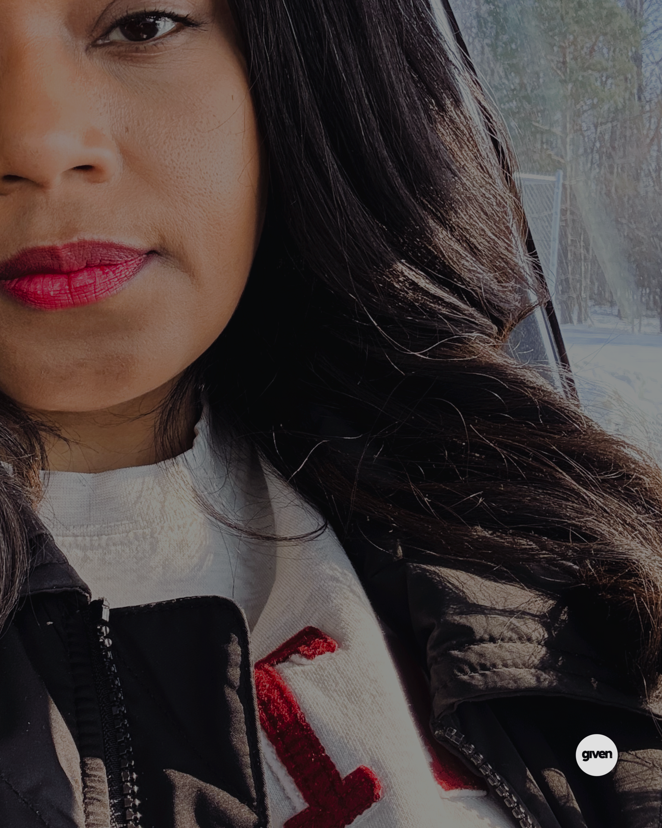 Close-up of a woman's face, showing her eye, nose, lips, and part of her dark brown hair. She's wearing a white shirt with red embroidery and a black jacket. The background features a blue sky and trees, indicating an outdoor setting.